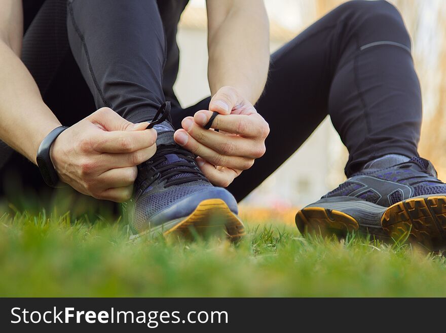 A Young Man In Black Clothes Is Tying The Laces On The Sneakers Close Up. Fitness Athlete Sitting On The Sports Field On The Grass