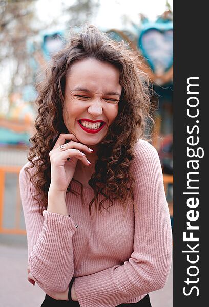 Girl chilling in amusement park in weekend morning. Laughing good-humoured female model with curly hair standing near carousel. Girl chilling in amusement park in weekend morning. Laughing good-humoured female model with curly hair standing near carousel.