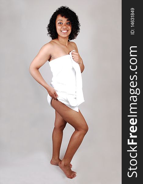 A young lovely Hispanic woman standing in the studio covert only in a white bath towel, bare feet and smiling into the camera, over light gray. A young lovely Hispanic woman standing in the studio covert only in a white bath towel, bare feet and smiling into the camera, over light gray.