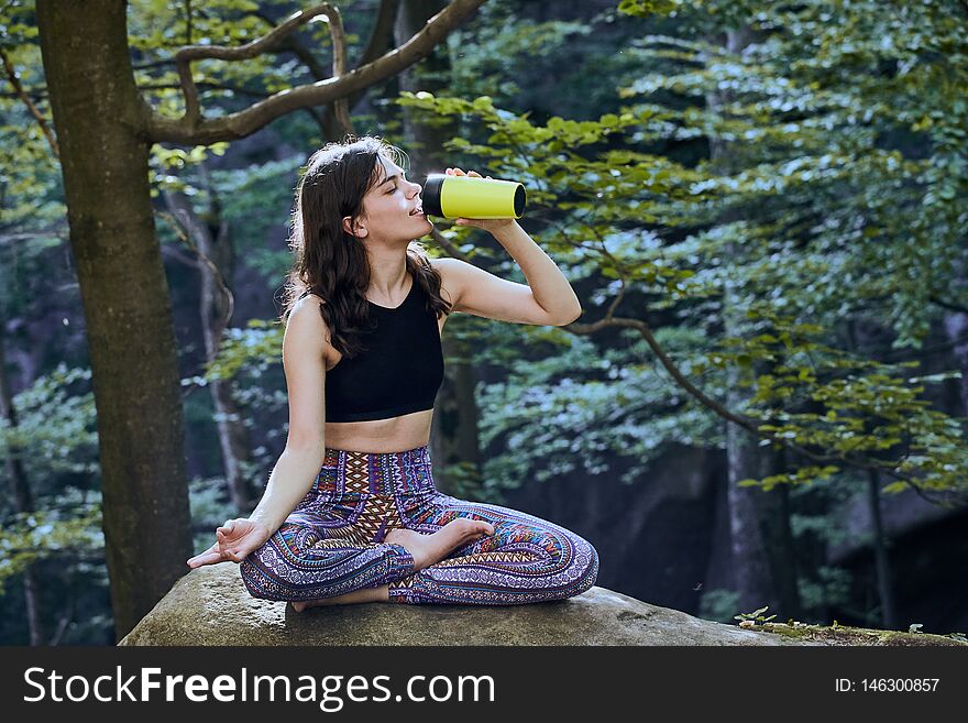 Healthy fitness girl sitting in lotus pose and drinking water from green bottle in forest