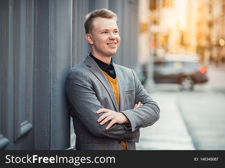 Handsome smiling young adult businessman standing outdoors near wall on city street.