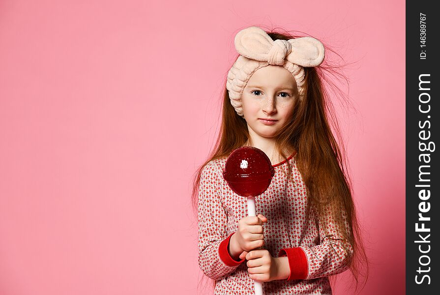 Funny red-haired baby girl in pajamas and dressing hair with a big red candy lollipop. Funny red-haired baby girl in pajamas and dressing hair with a big red candy lollipop