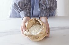 Paper Bag With White Flour. Woman Standing Next To The Kitchen Table And Holding Paper Bag, Flour In It Stock Photo