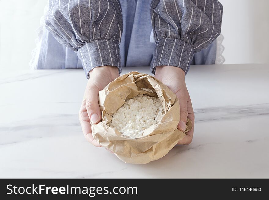 Paper bag with white flour. Woman standing next to the kitchen table and holding paper bag, flour in it