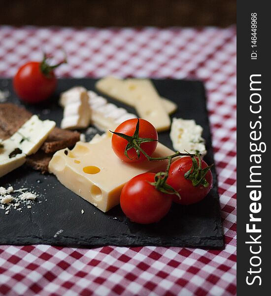 Delicious plate of french cheese served with fresh ripe tomatoes and bread on a black charcoal board. Red and white tablecloth for a picnic mood. Delicious plate of french cheese served with fresh ripe tomatoes and bread on a black charcoal board. Red and white tablecloth for a picnic mood