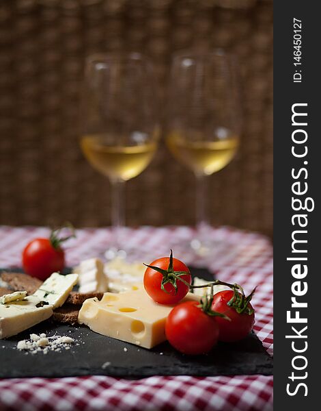 Delicious plate of french cheese served with fresh ripe tomatoes and bread on a black charcoal board. Red and white tablecloth for a picnic mood. Two glasses of white wine on background. Delicious plate of french cheese served with fresh ripe tomatoes and bread on a black charcoal board. Red and white tablecloth for a picnic mood. Two glasses of white wine on background