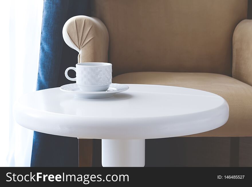 White tea cup and saucer on a round white table against a beige armchair
