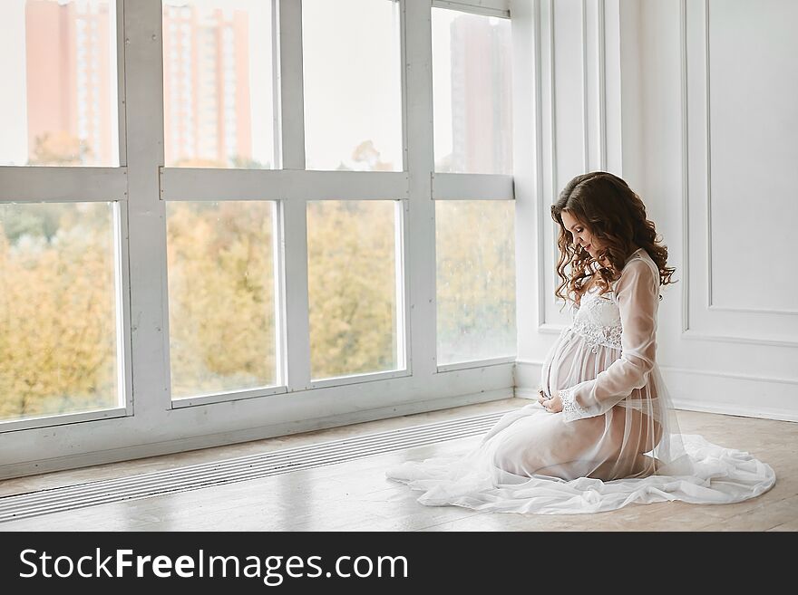 Beautiful young pregnant woman with fashionable hairstyle in a stylish peignoir smiling and posing near the big window at light interior.