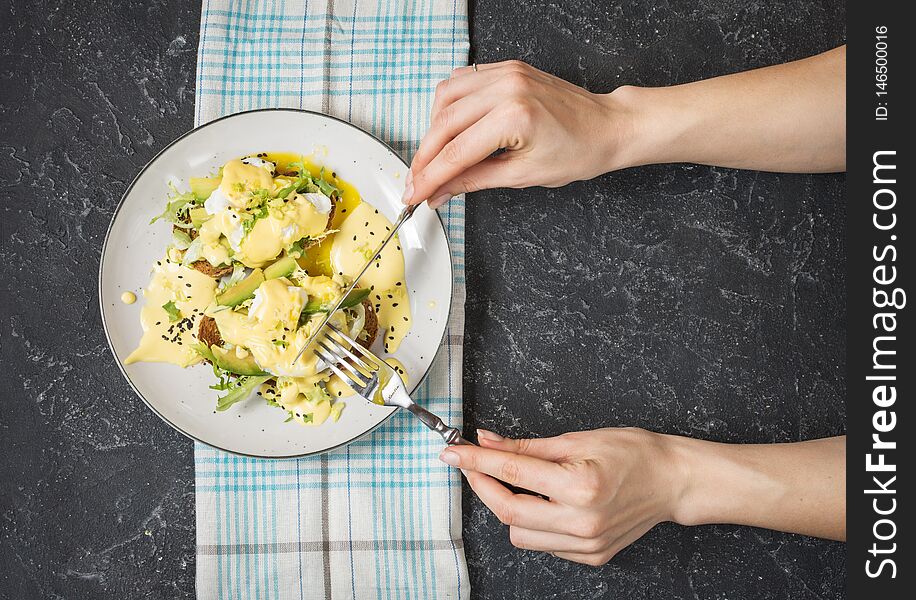 Female Hand Hold Fork And Knife. Eggs Benedict With Salad And Avocado On Stone Background