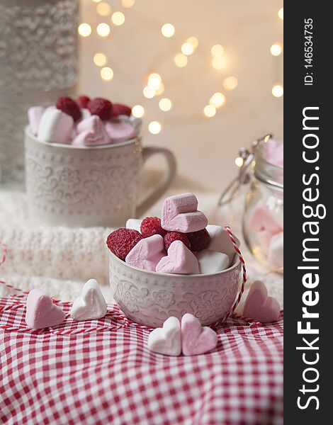 Home saint valentine`s day party: heart shaped white and pink marshmallow served with fresh raspberry. Table decorated with red tablecloth and white knitted rud and lights on a background. Home saint valentine`s day party: heart shaped white and pink marshmallow served with fresh raspberry. Table decorated with red tablecloth and white knitted rud and lights on a background