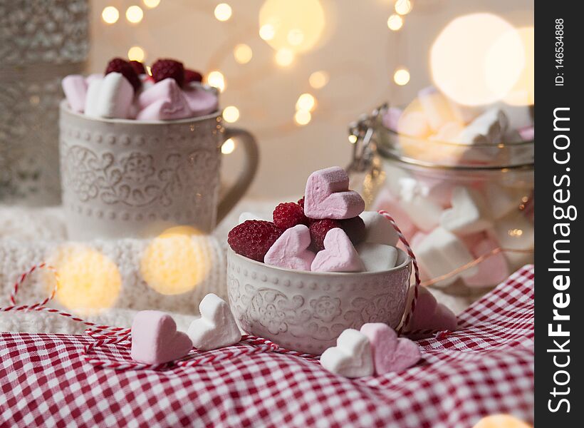 Home saint valentine`s day party: heart shaped white and pink marshmallow served with fresh raspberry. Table decorated with red tablecloth and white knitted rud and lights on a background. Home saint valentine`s day party: heart shaped white and pink marshmallow served with fresh raspberry. Table decorated with red tablecloth and white knitted rud and lights on a background