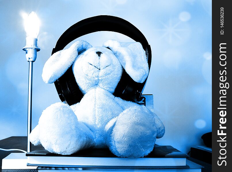 Rabbit plush toy with wireless headphones sitting on books enjoying music. Soft blue bokeh effect
