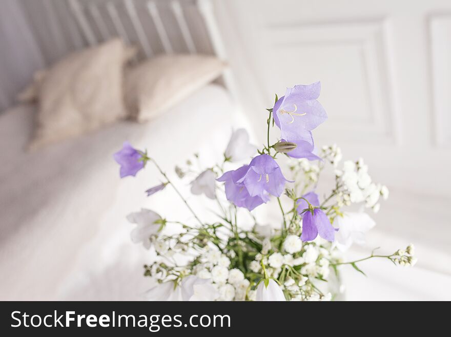 Glass vase with lilac and white floweers  in light cozy bedroom interior. White wall, bed with white linen, light blanket or plaid and pillows
