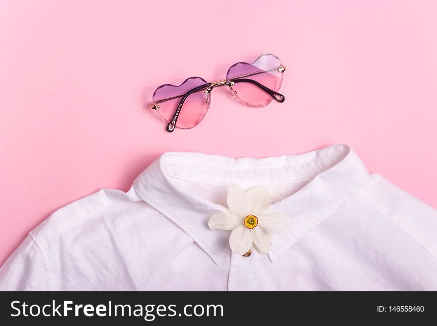 Collarless white shirt with stylish heart-shaped glasses and a spring flower on a pink background. Top view, flat lay. Collarless white shirt with stylish heart-shaped glasses and a spring flower on a pink background. Top view, flat lay