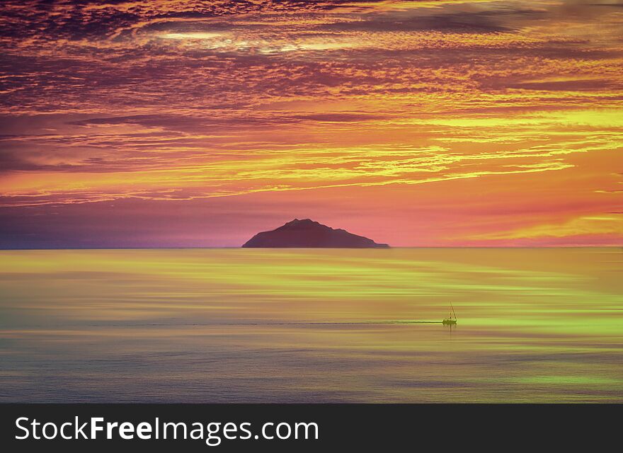 A fiery sunset with a boat that travels in front of an island and a sky that is reflected in the sea