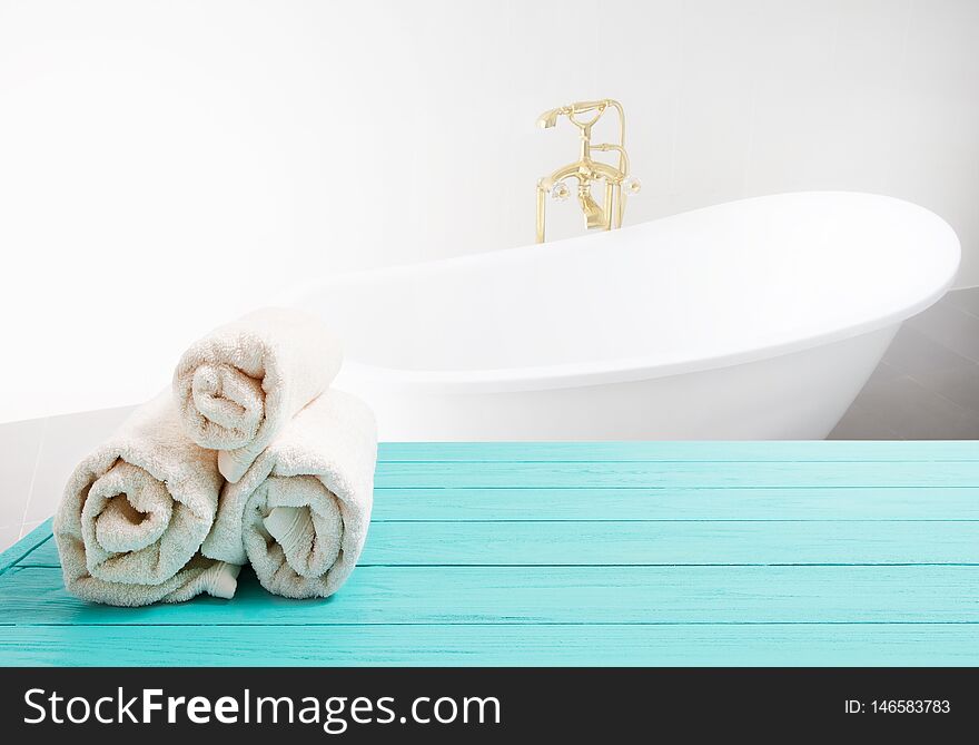 Blue wooden shelf with rolled towels on bathroom background. Wood desk table with top view and copy space near white bath at hotel. Room service and shower spa concept. Selective focus