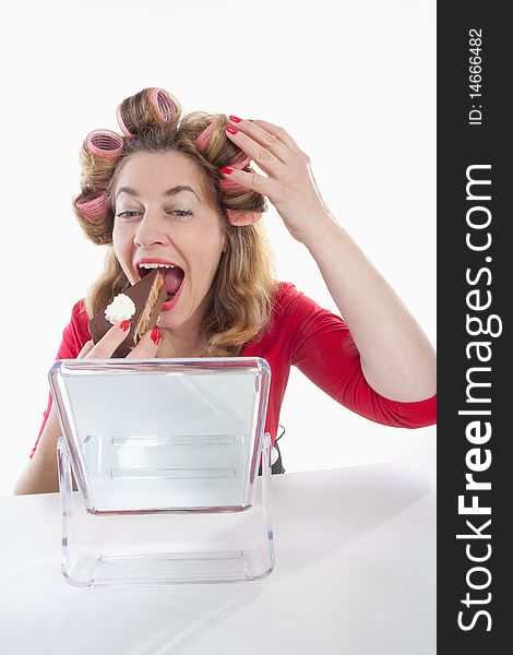 Middle-aged woman with hair rollers eating cake looking at mirror