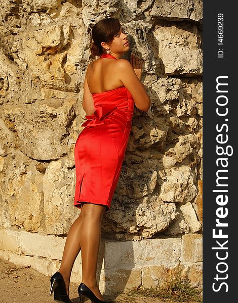 The beautiful girl in a red dress costs at a stone wall. The beautiful girl in a red dress costs at a stone wall