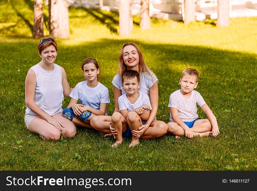 Happy women and children sit on the green grass and look at the camera. Big happy family, two mothers and three children in white t-shirts and shorts. Summer day