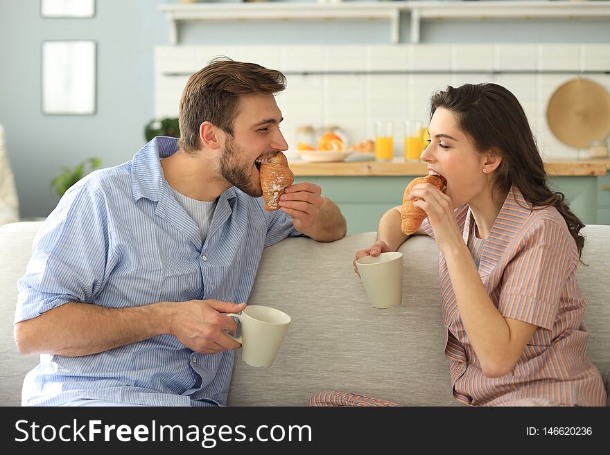 Happy Young Couple In Pajamas In Kitchen Having Breakfast, Feeding Each Other A Croissant