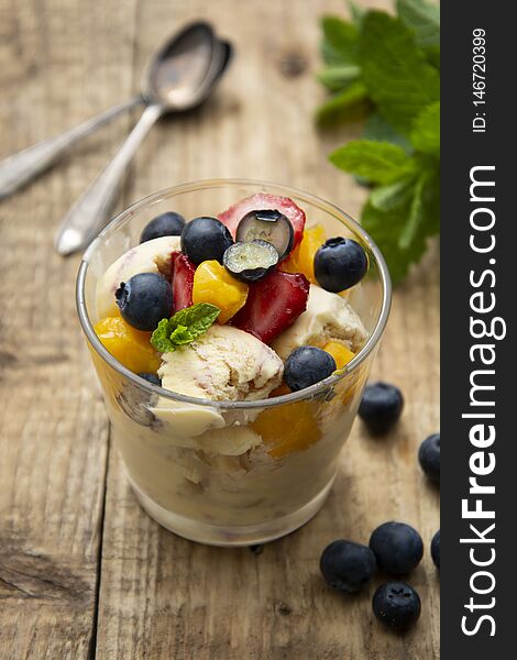 Vanilla ice cream with fruits - blueberries, strawberries, mango and mint, summer refreshing dessert. Wooden background. Glass with vanilla ice cream