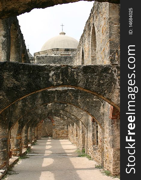 Looking down exterior hallway of Spanish mission with domed roof and cross in background.