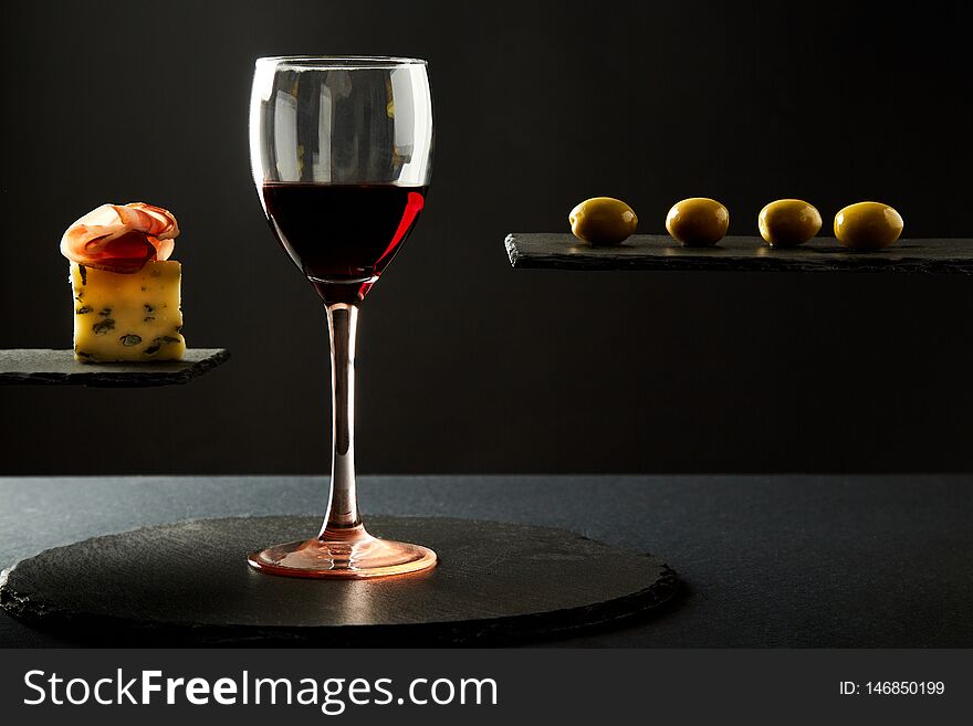 Composition of red wine in glass and pieces of prosciutto and cheese and olives on black background.