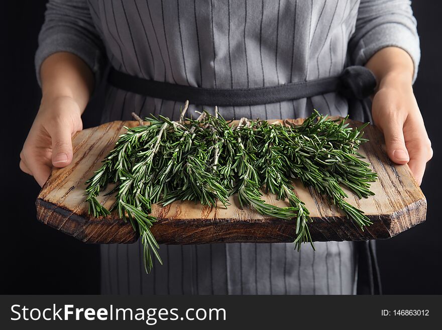 Woman holding wooden board with fresh rosemary twigs on black background, closeup