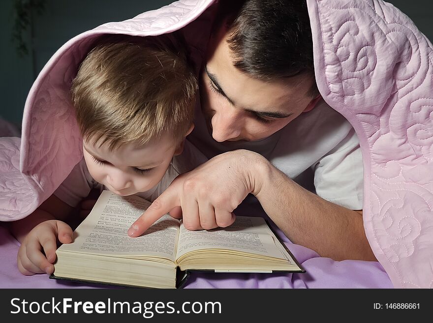 Dad teaches his little son to read a book hiding under the blanket.
