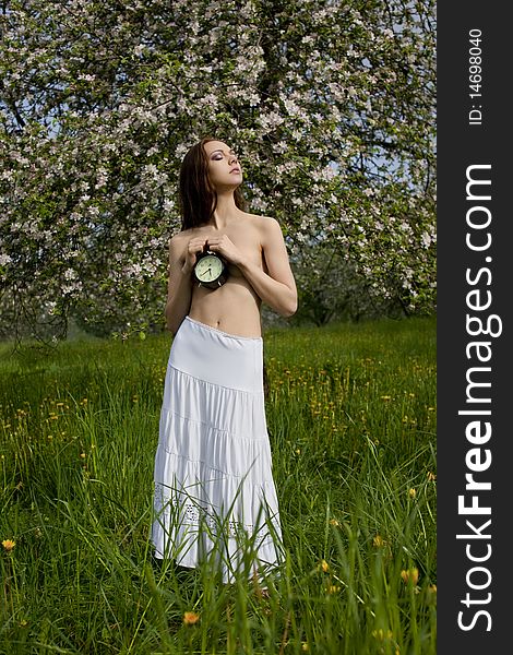 Young girl in a white skirt with an alarm clock in the hands near blooming apple tree. Young girl in a white skirt with an alarm clock in the hands near blooming apple tree