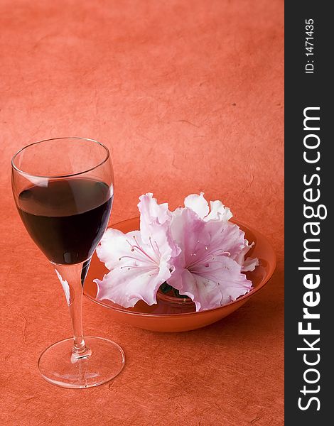 Studio shot of red wine in a wine glass with red glass bowl of a few azalea flowers. Shot against a red textured background. Studio shot of red wine in a wine glass with red glass bowl of a few azalea flowers. Shot against a red textured background.