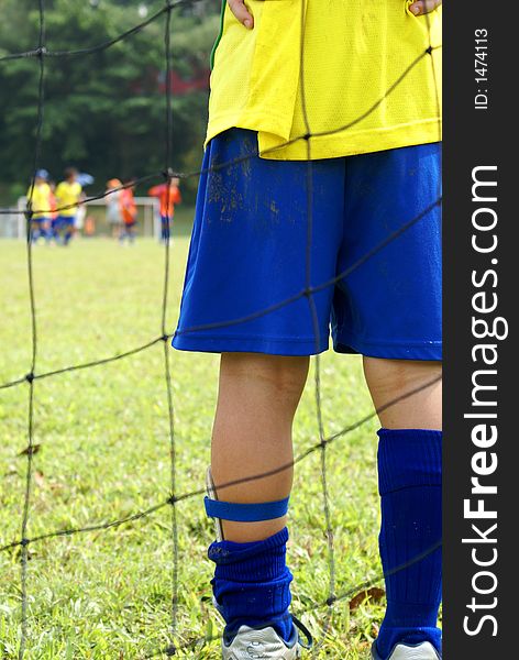 A picture of a young goalkeeper's legs. Netting in the foreground while other children is at the other end of the field. Yellow and blue. A picture of a young goalkeeper's legs. Netting in the foreground while other children is at the other end of the field. Yellow and blue.