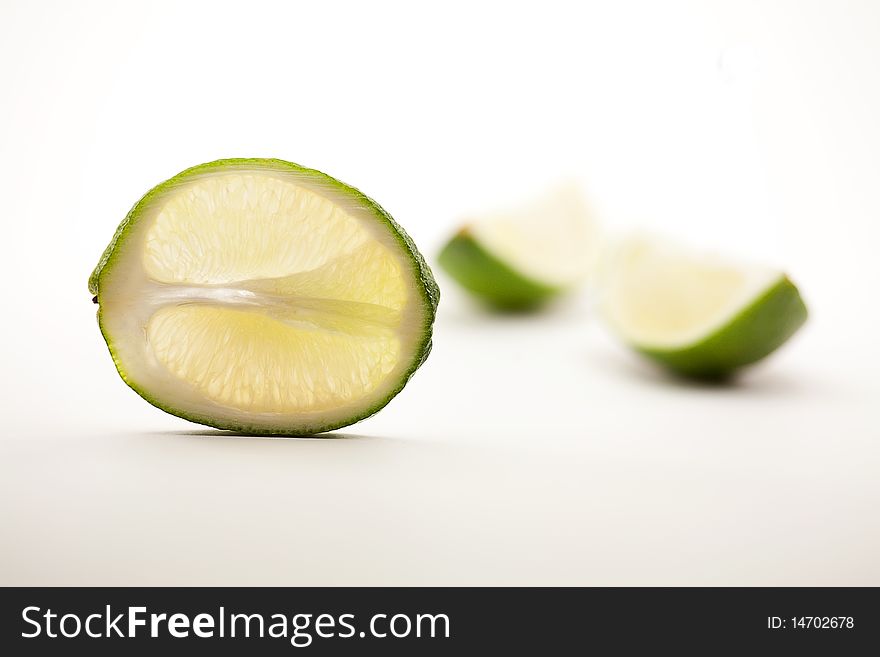 One lime slice standing vertically upright with two quarter lime segments out of focus on white background. One lime slice standing vertically upright with two quarter lime segments out of focus on white background