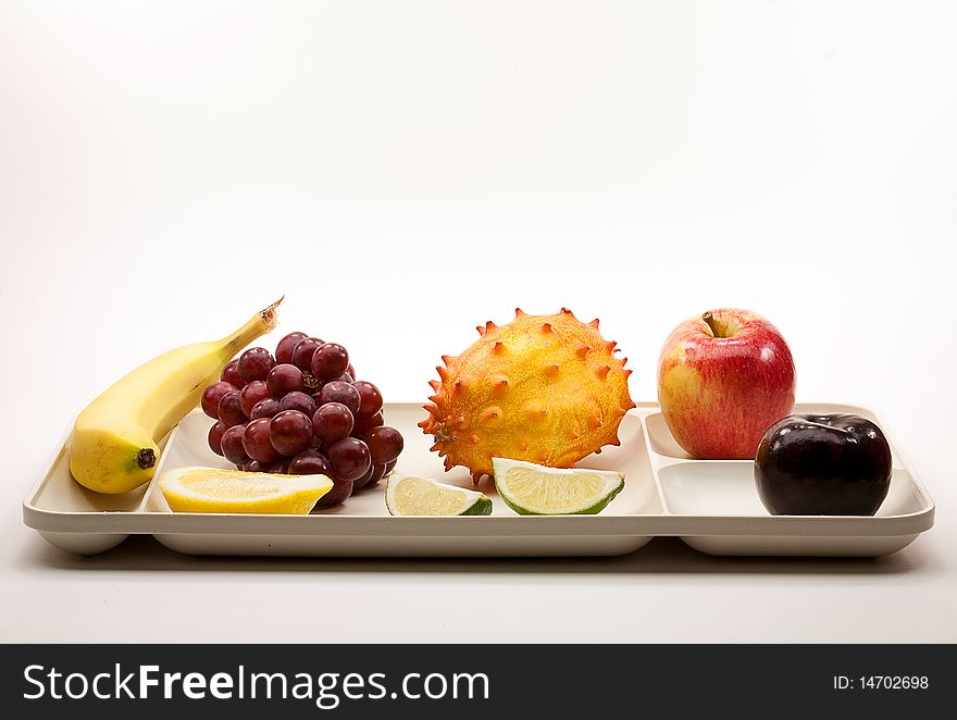 Banana, grapes, lemon, lime, horned melon, apple and plum resting on a plastic picnic serving tray with white background. Banana, grapes, lemon, lime, horned melon, apple and plum resting on a plastic picnic serving tray with white background.