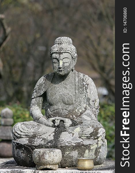 A stone statue of Buddha against a natural setting. This statue is old and is now covered with lichens. It's in a meditative pose with a stone and bronze pot in front of him. A stone statue of Buddha against a natural setting. This statue is old and is now covered with lichens. It's in a meditative pose with a stone and bronze pot in front of him.