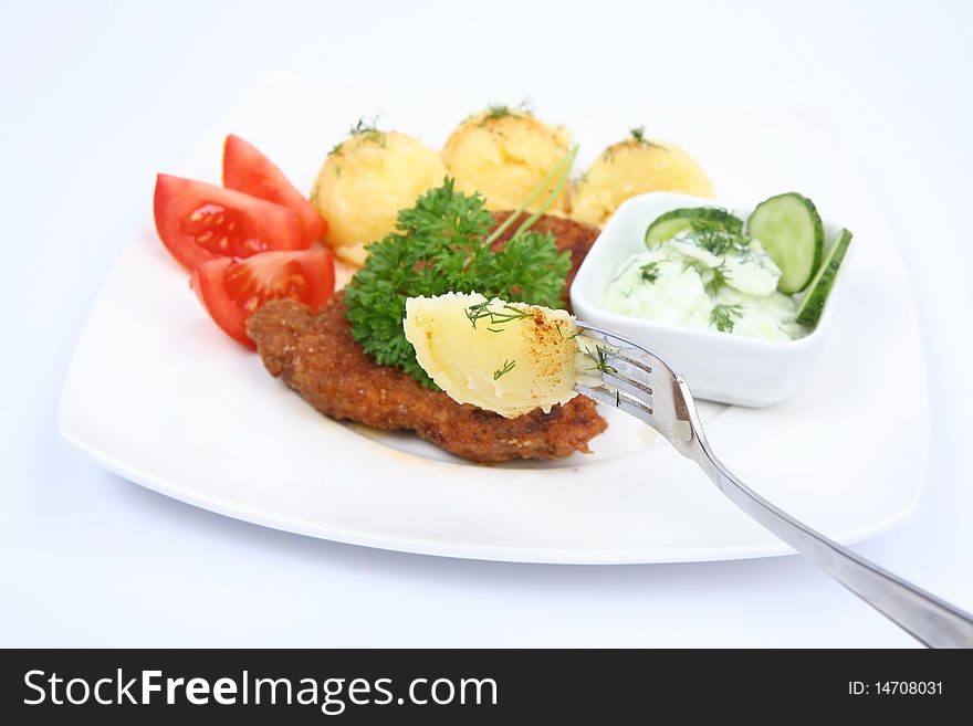 Pork chop (covered in batter and breadcrumbs), mashed potatoes-some of it on a fork, cucumber salad in a bowl and tomato pieces decorated with dill and parsley on a plate with a on white background. Pork chop (covered in batter and breadcrumbs), mashed potatoes-some of it on a fork, cucumber salad in a bowl and tomato pieces decorated with dill and parsley on a plate with a on white background