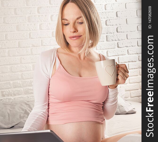 Happy smiling pregnant woman is sitting in bed and looks something in the tablet and drinks morning coffee . Online Shopping. Happy smiling pregnant woman is sitting in bed and looks something in the tablet and drinks morning coffee . Online Shopping.