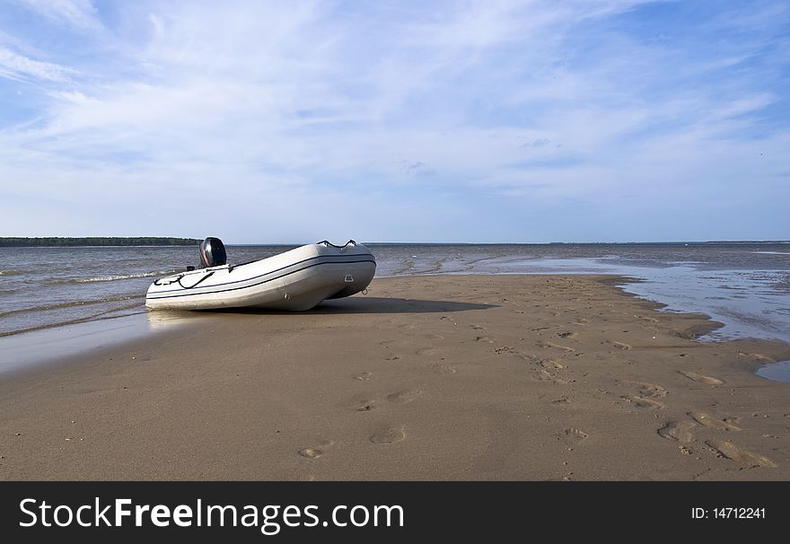 Inflatable fishing boat with an engine standing on a deserted beach on the background of blue sky with clouds. Landscape.