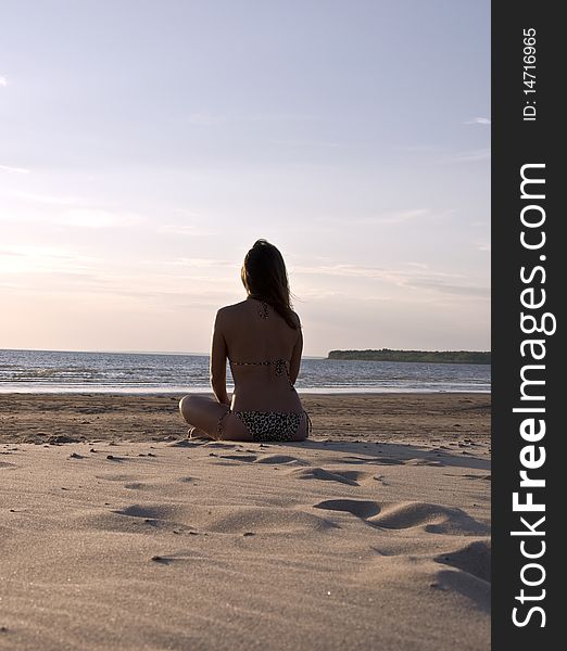 Young girl sitting on a deserted beach back to the camera and admires the sunset. Landscape.