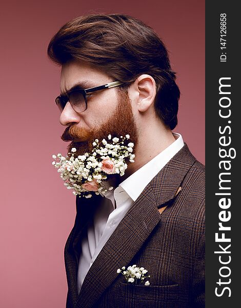 Profile portrait of a fashionable bearded man with flowers in beard, isolated on a dark pink background.