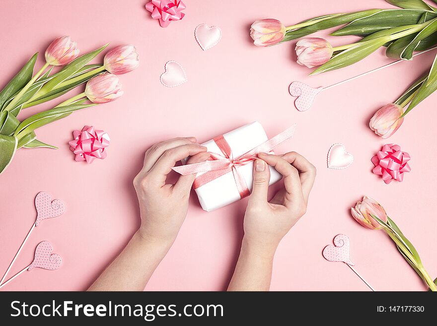 Women`s hands decorate gift box surrounded tulip flowers and hearts on pink pastel background