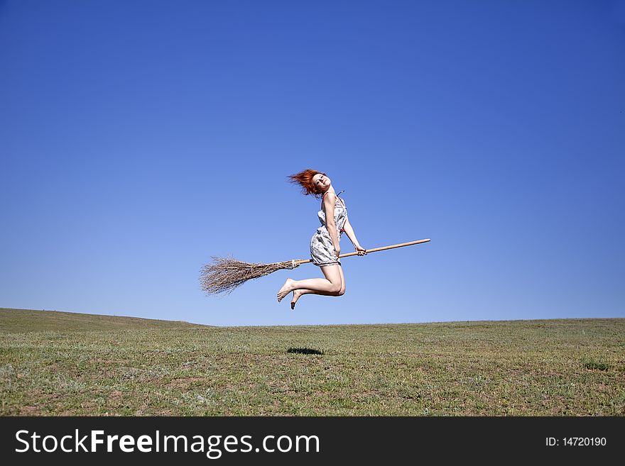 Young red-haired witch on broom flying over green grass field.