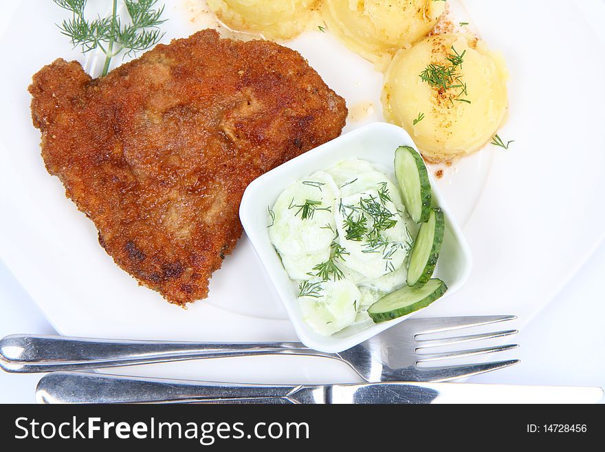 Pork chop (covered in batter and breadcrumbs), mashed potatoes and cucumber salad in a bowl decorated with dill on a plate with a fork and a knife on white background. Pork chop (covered in batter and breadcrumbs), mashed potatoes and cucumber salad in a bowl decorated with dill on a plate with a fork and a knife on white background