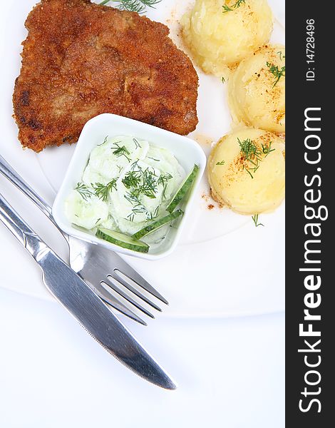 Pork chop (covered in batter and breadcrumbs), mashed potatoes and cucumber salad in a bowl decorated with dill on a plate with a fork and a knife on white background. Pork chop (covered in batter and breadcrumbs), mashed potatoes and cucumber salad in a bowl decorated with dill on a plate with a fork and a knife on white background