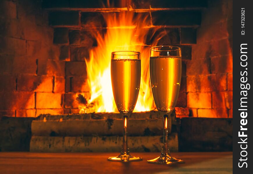 Two glasses with champagne on a wooden table near the fireplace with burning birch wood.