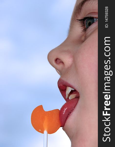 Young blue eyed girl licking an orange lollipop into a heart shape against a blue sky. Young blue eyed girl licking an orange lollipop into a heart shape against a blue sky