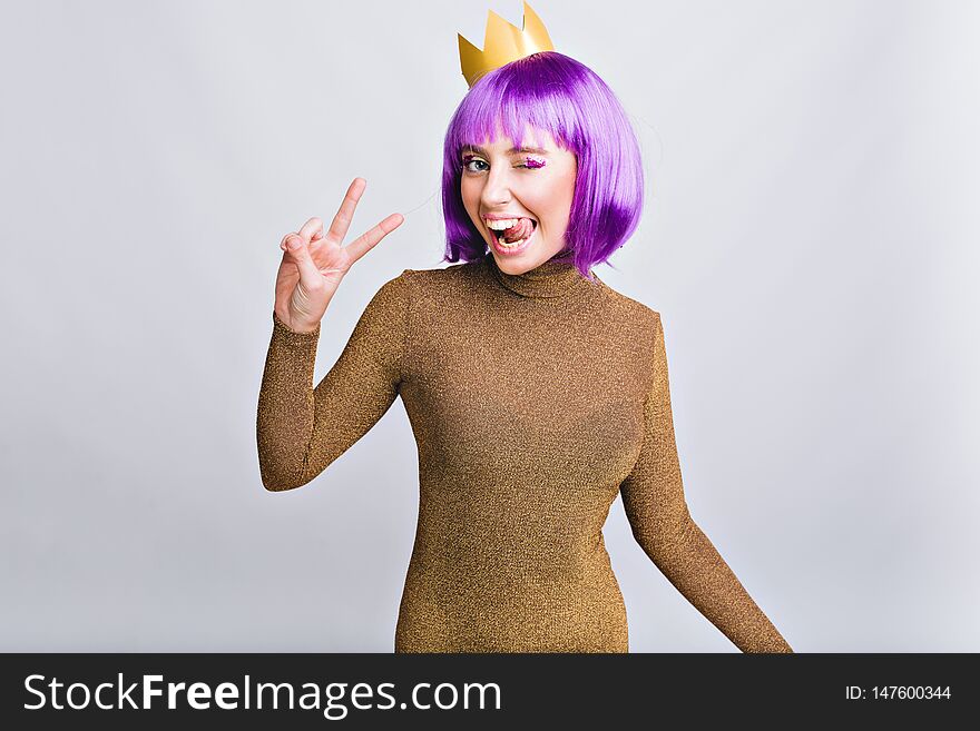 Portrait pretty girl with gold crown having fun in studio. She wears violet haircut, shows tongue and looks happy to