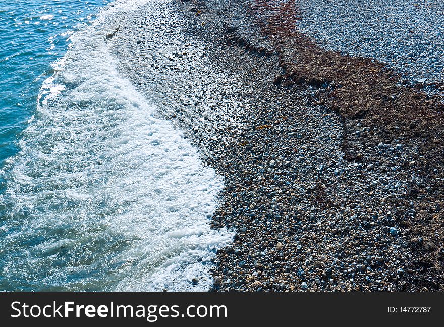 A background texture of turquoise sea, waves, and a shingle beach. Pebbles of different sizes and seaweed lie on the beach. A background texture of turquoise sea, waves, and a shingle beach. Pebbles of different sizes and seaweed lie on the beach.
