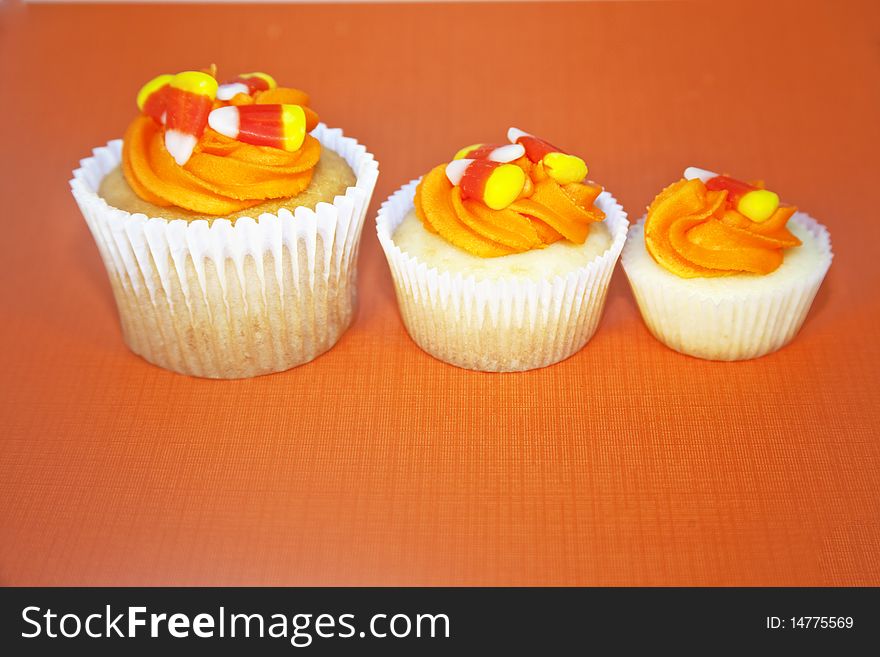 Three different size vanilla cupcakes decorated with orange butter cream frosting and candy corn - all on an orange background. Three different size vanilla cupcakes decorated with orange butter cream frosting and candy corn - all on an orange background.