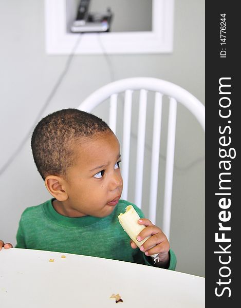 Young child sitting at table with banana in hand eating while looking away. Young child sitting at table with banana in hand eating while looking away